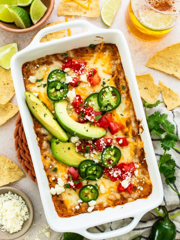 Bean dip topped with melted cheese and garnished with diced tomatoes, cotija cheese, sliced peppers, and avocados.