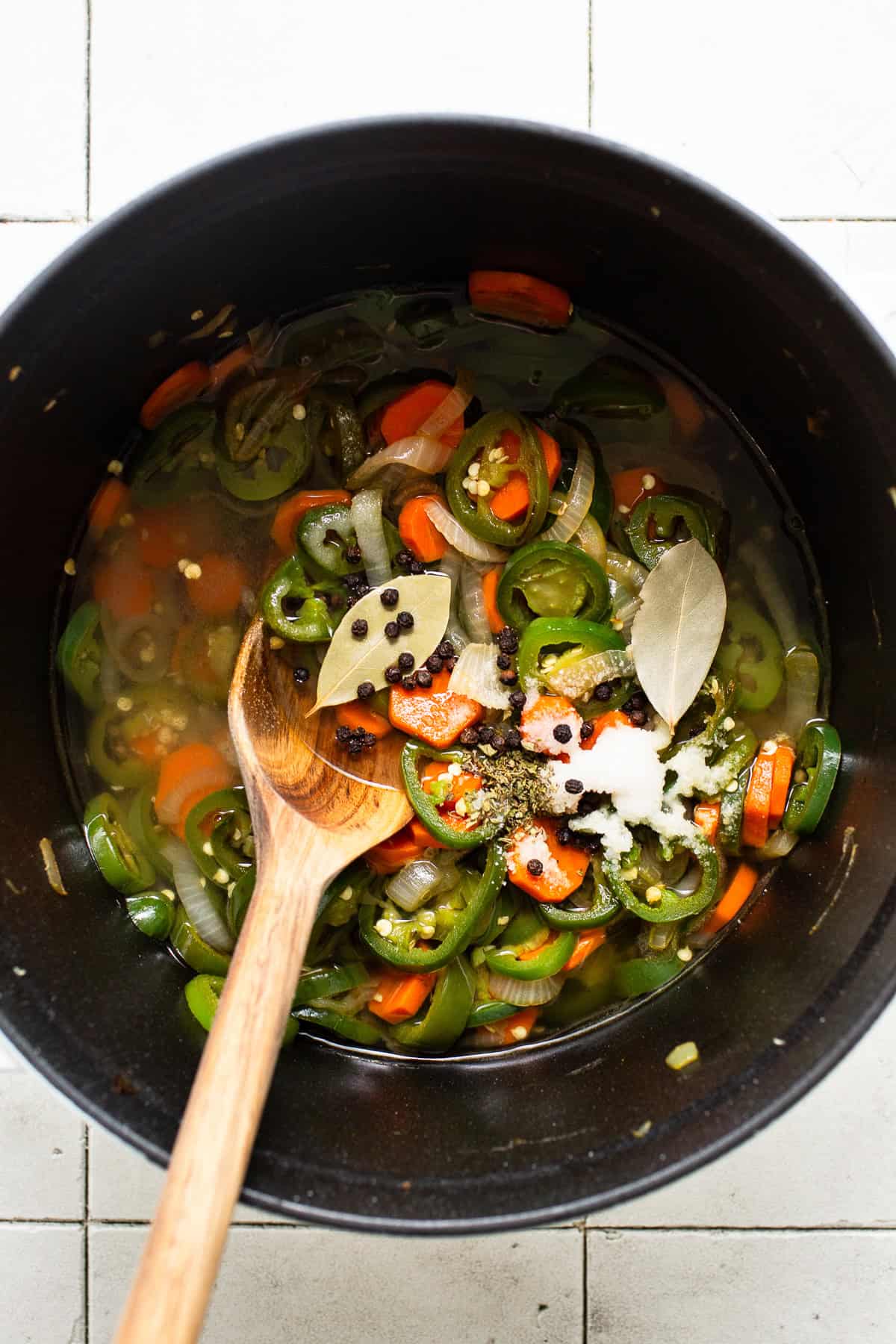 Jalapeños, onions, and carrots being pickled in a pot with lime juice, bay leaves, black peppercorns, salt, and oregano.