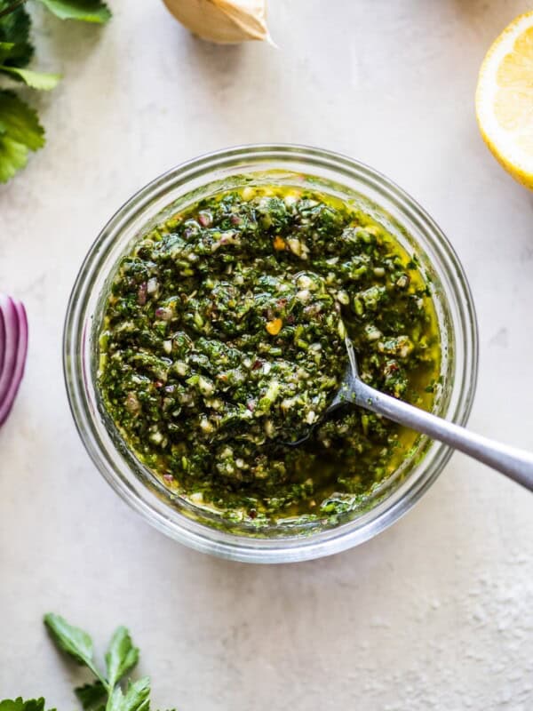 Chimichurri sauce in a clear bowl surrounded by parsley, red onions, cilantro, garlic and lemon juice.