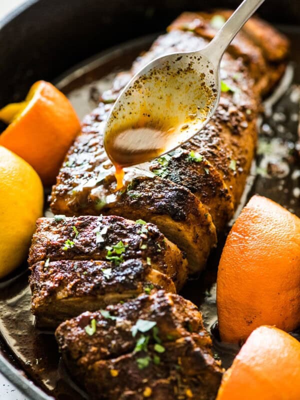 A spoon pouring citrus juices over roasted pork tenderloin in a black cast iron skillet.