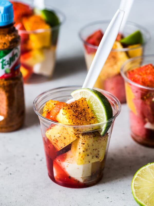 Mexican Fruit Cups on a table topped with chile lime seasoning, chamoy and a lime wedge.