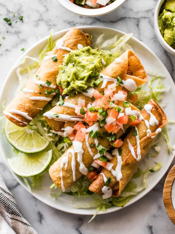 Chicken taquitos on a plate garnished with Mexican crema, lettuce, guacamole, and pico de gallo.