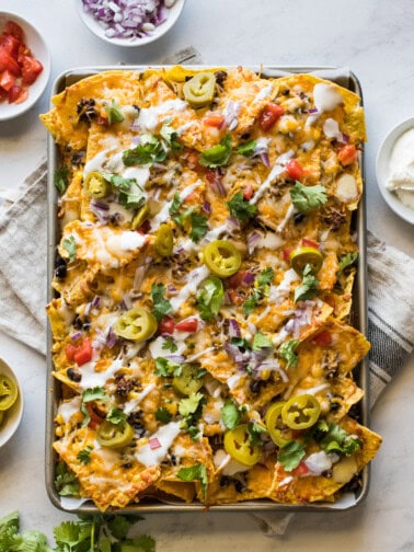 Sheet pan nachos topped with sour cream, pickled jalapenos, tomatoes, and cilantro.