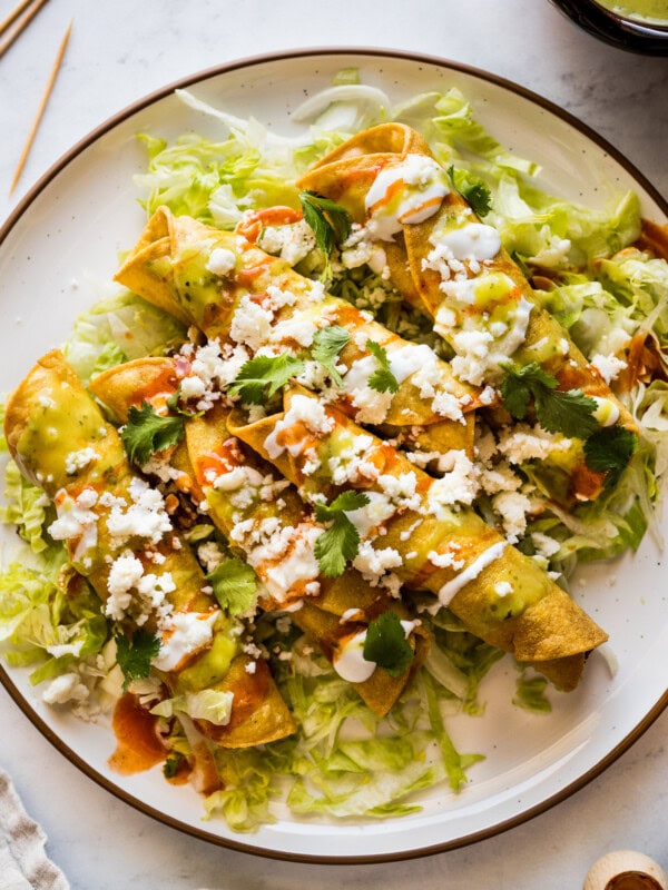 Flautas on a plate topped with crema, hot sauce, salsa verde, lettuce, cilantro, and cotija cheese.