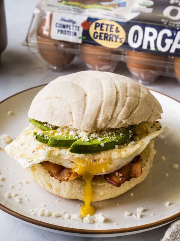 A concha breakfast sandwich on a plate made from bacon, a fried egg, avocado, cotija cheese, and a maple conchas.