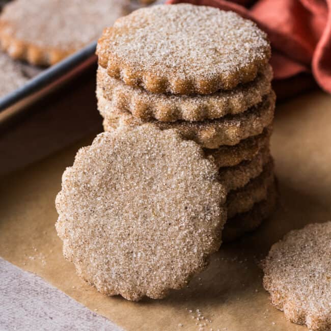 Biscochitos covered in cinnamon sugar stacked on top of one another on a piece of parchment paper.