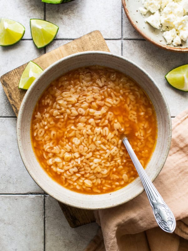 A bowl of sopa de letras on a table with limes, cheese, and avocado on the side.