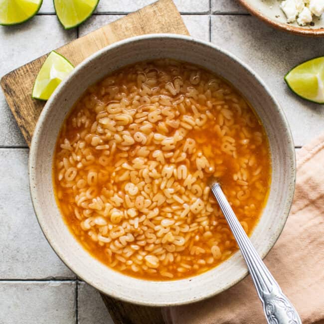 A bowl of sopa de letras on a table with limes, cheese, and avocado on the side.