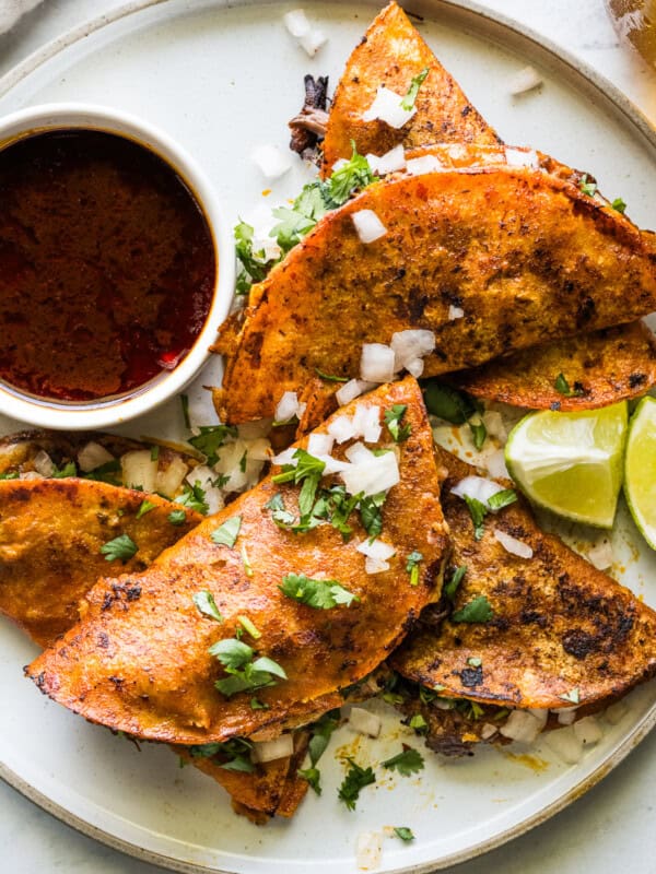 Birria tacos on a plate served with birria consomé and topped with diced white onion, cilantro, and limes.