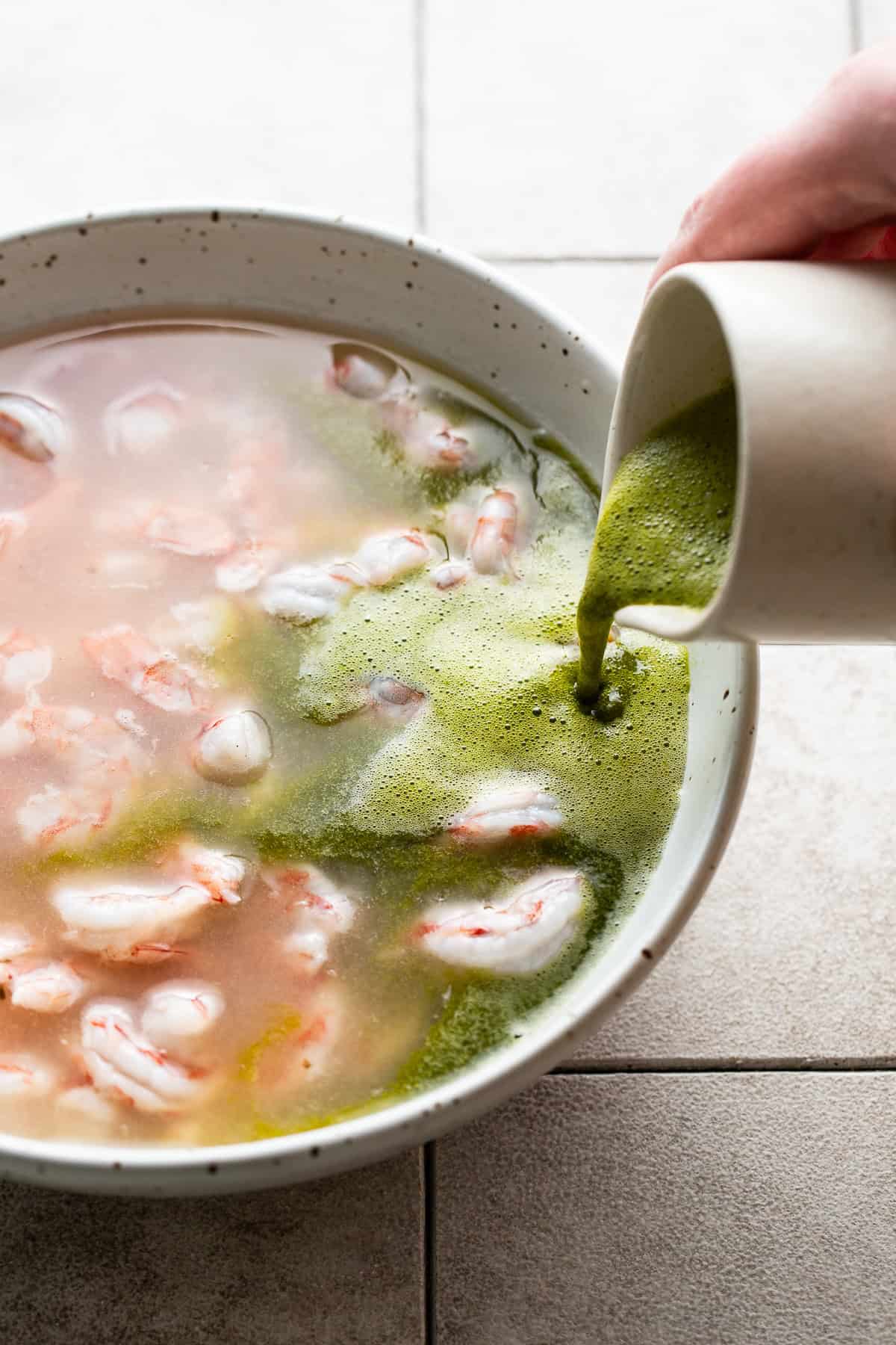 The blended cilantro, serrano, and lime juice mixture being poured into a bowl of shrimp aguachile.
