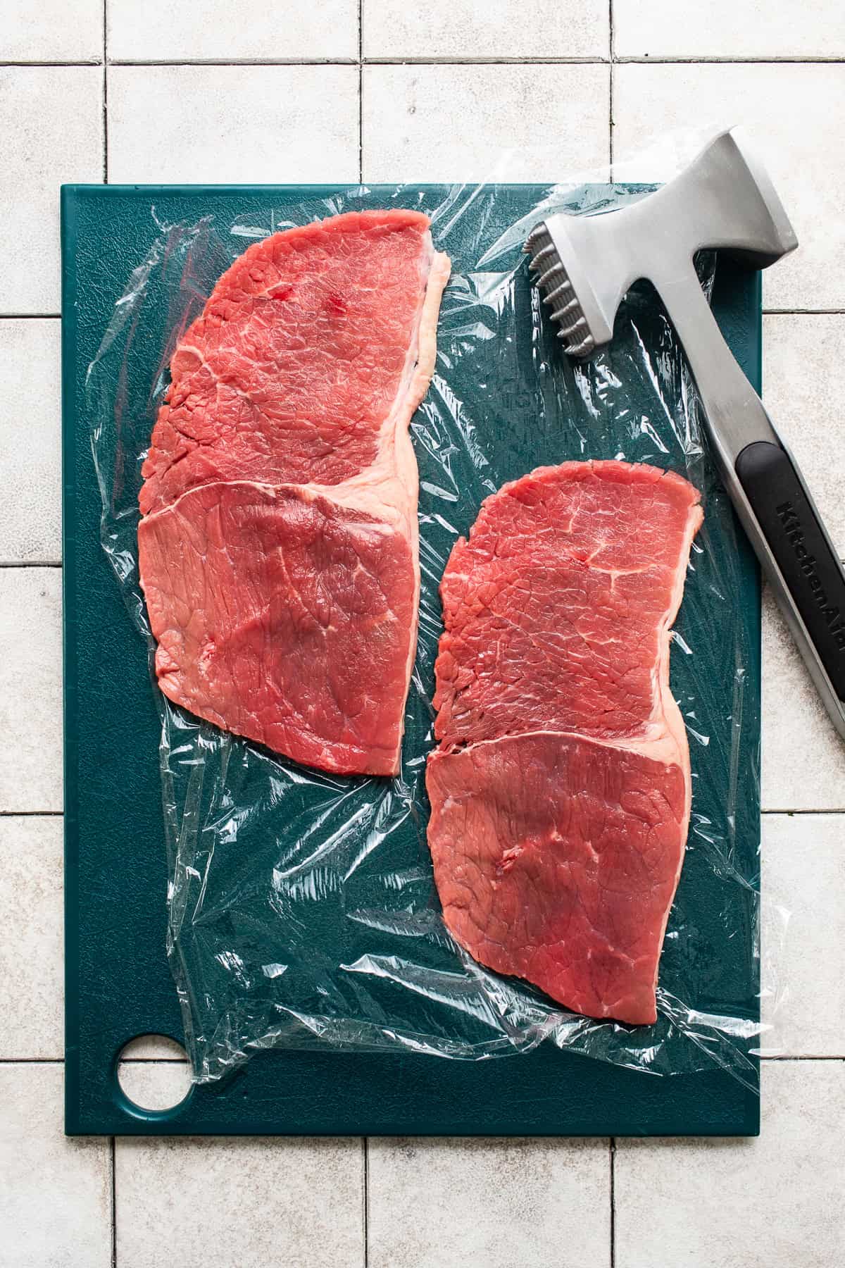 Thinly sliced steak that has been pounded with a meat mallet on a cutting board lined with plastic wrap.