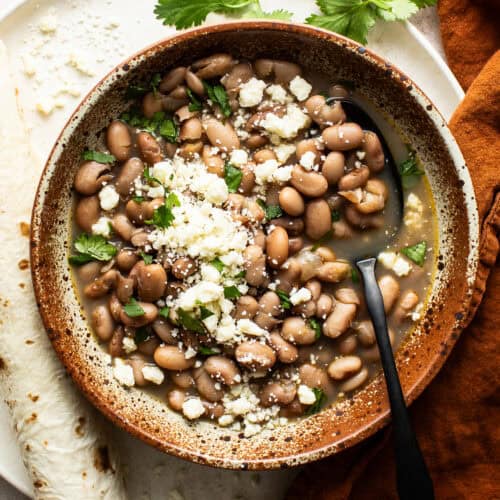 Cooked pinto beans in a bowl topped with cilantro, cotija cheese, and served with flour tortillas
