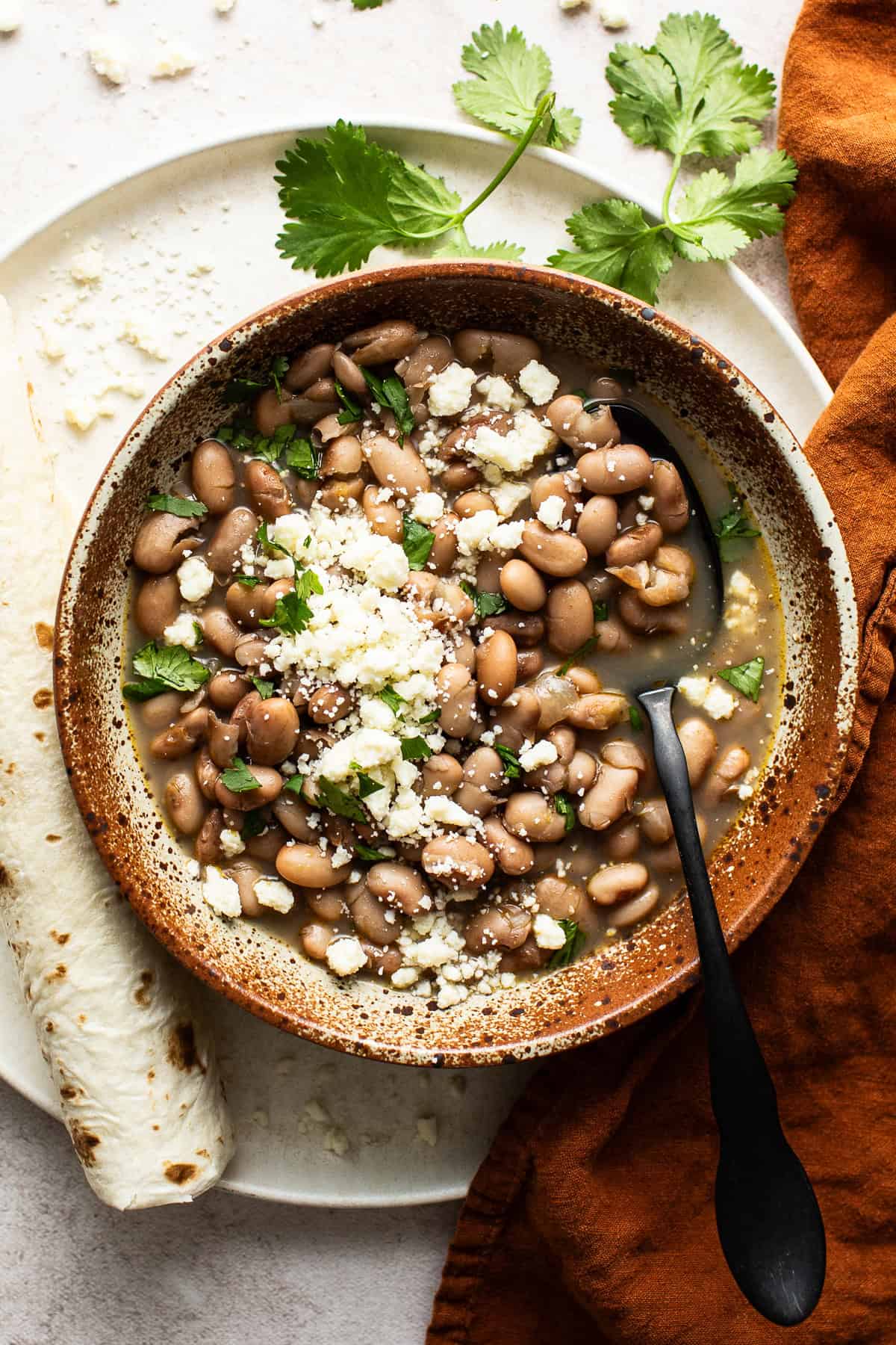 Cooked pinto beans in a bowl topped with cilantro, queso fresco, and served with flour tortillas