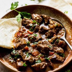 a bowl of carne guisada with garnish and a flour tortilla, ready to eat.