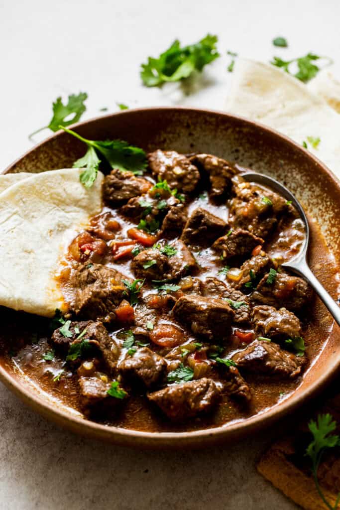 A bowl of carne guisada with garnish and a flour tortilla, ready to eat.