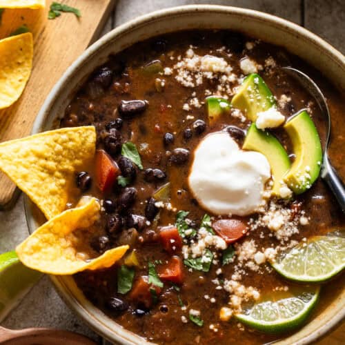 Vegetarian black bean soup in a bowl topped with avocados, cilantro, and limes.