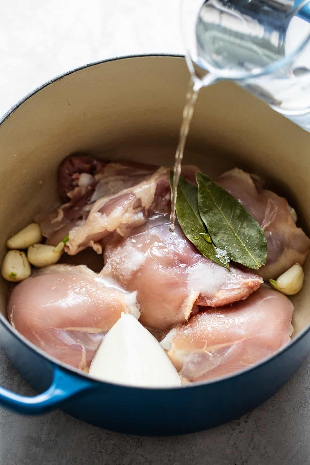 Water being poured into a pot with chicken thighs, onion, garlic, and bay leaves to make Mexican caldo tlalpeño.