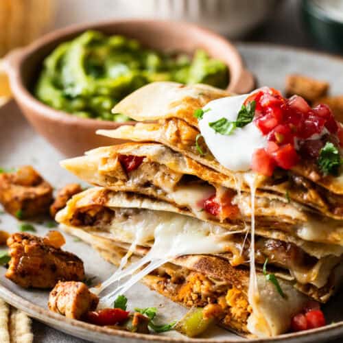 Chicken fajita quesadillas stacked on a plate with sour cream, lettuce, and tomatoes on top.