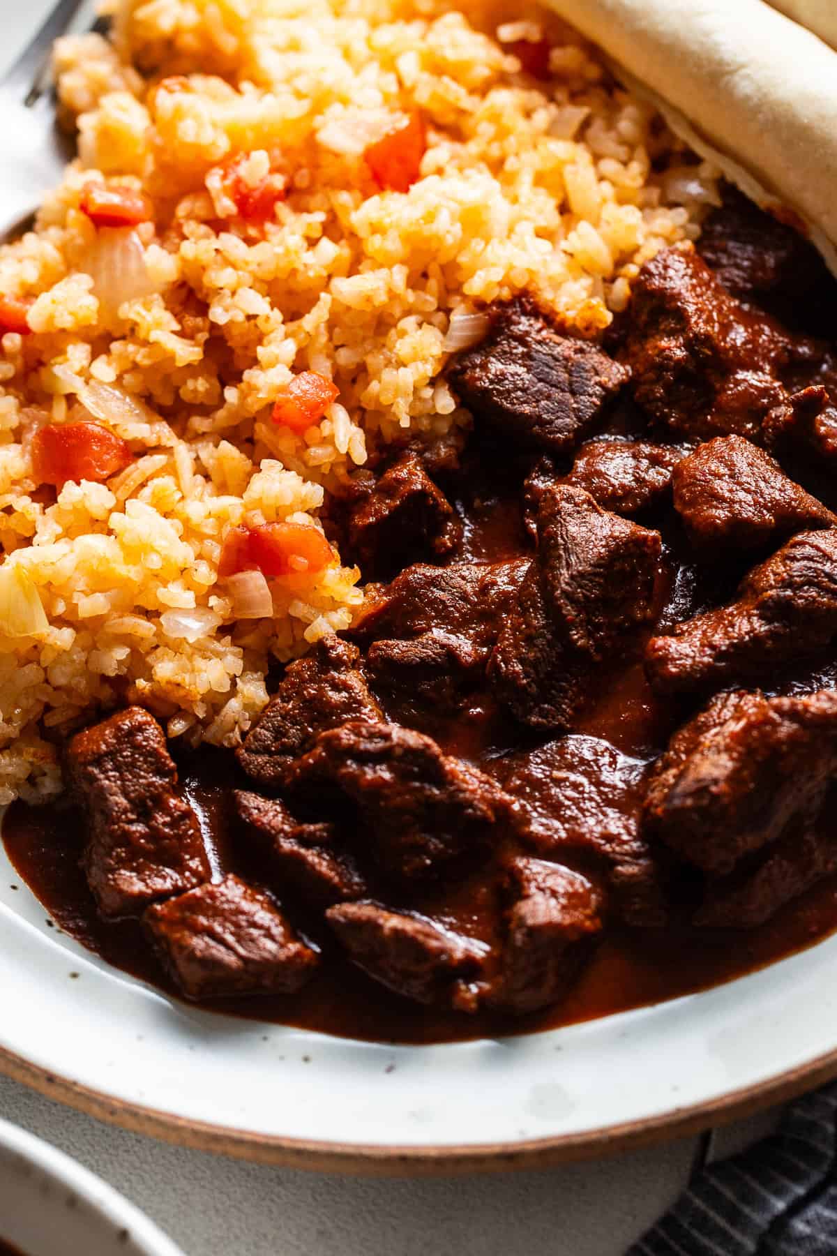 Traditional chile colorado made from tender pieces of beef in a red chile sauce on a plate next to red Mexican rice.