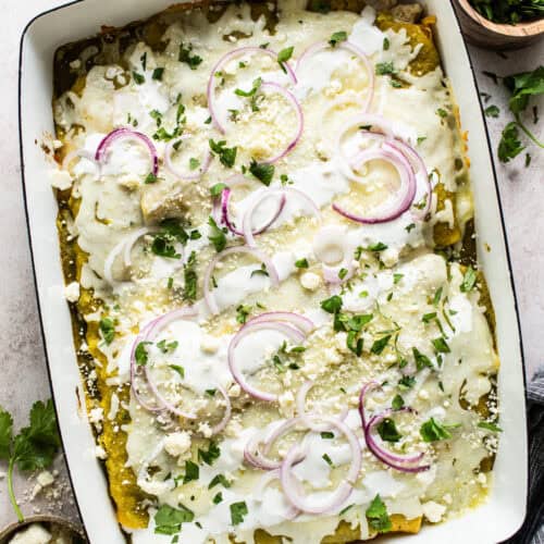 Enchiladas Verdes in a baking dish topped with cilantro, red onion, and Mexican crema or sour cream.