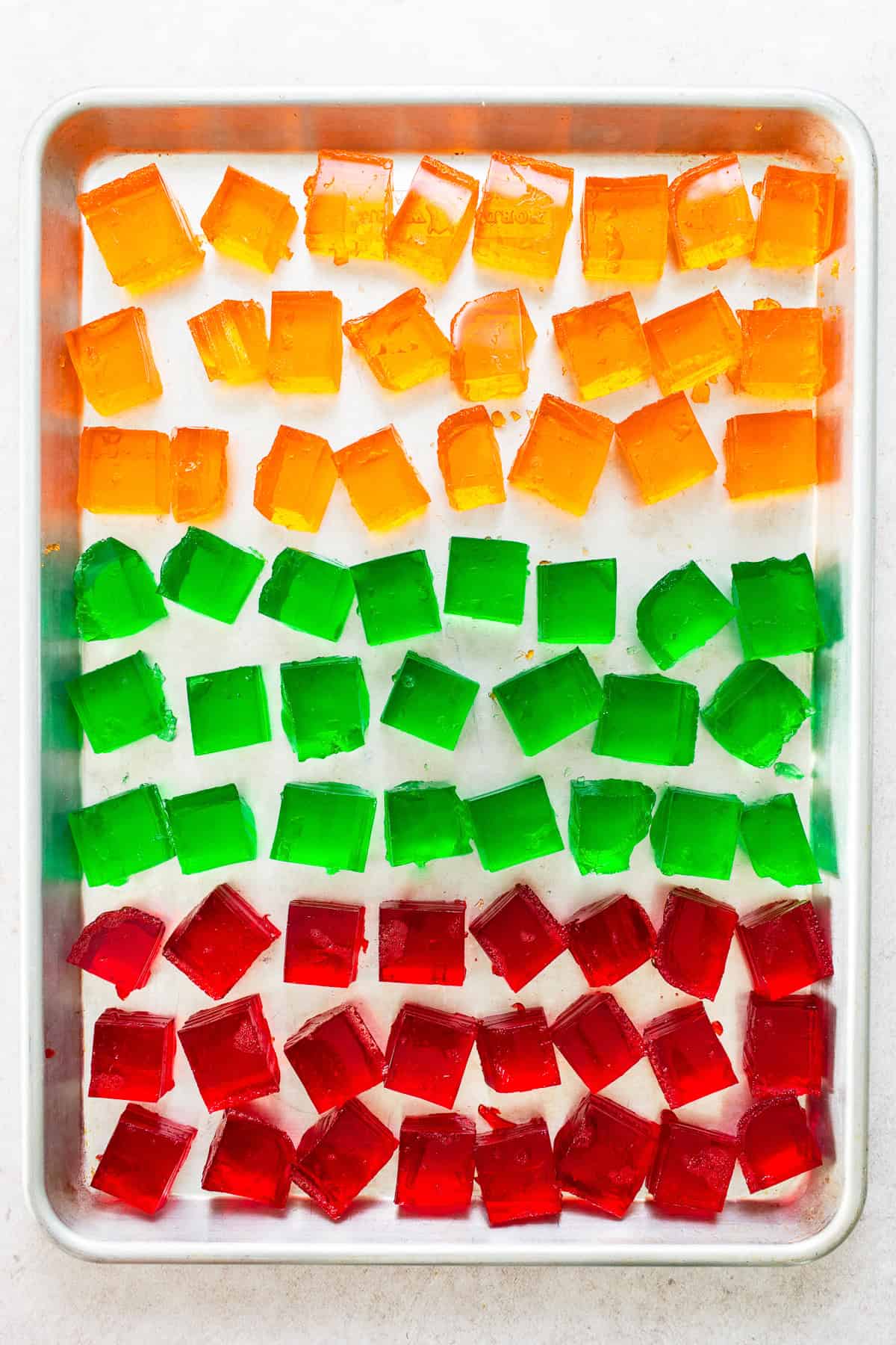 Orange, green, and red cubes of jello sliced into squares and placed on a baking sheet to assemble gelatina de mosaico.