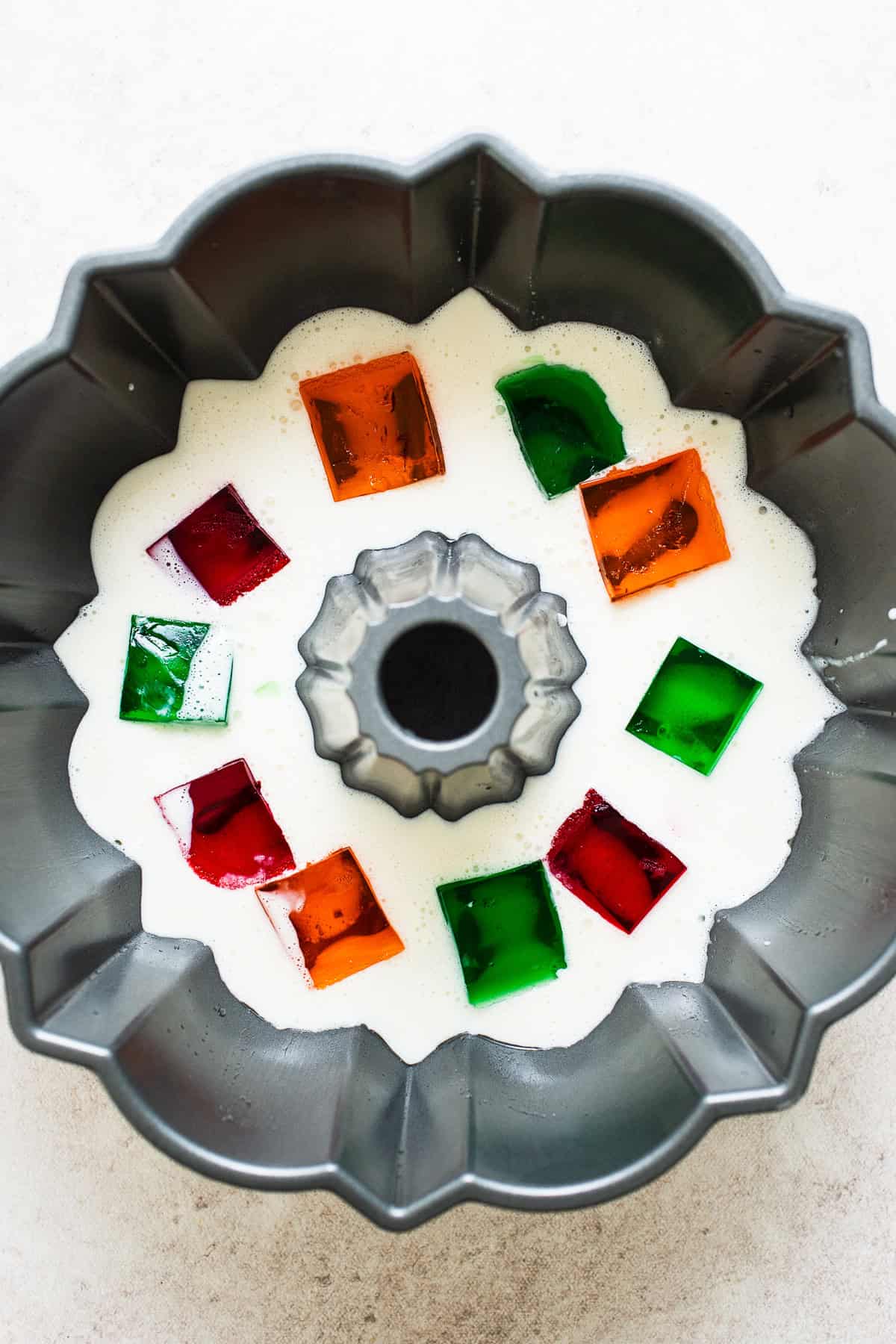 Gelatina de mosaico being assembled in a bundt pan with white milk gelatina and cubes of different colored flavored jellos.