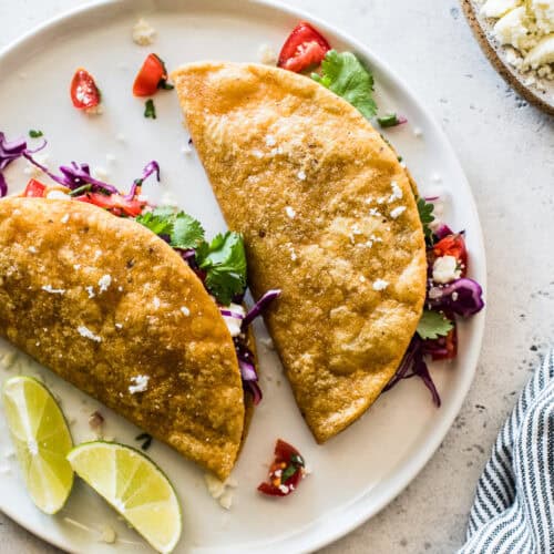 A plate of two crispy potato tacos, garnished with limes and ready to be served.