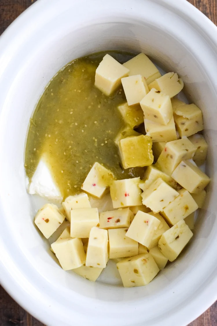 A crockpot filled with salsa verde, cheese cubes, and cream cheese to make salsa verde queso in the slow cooker.