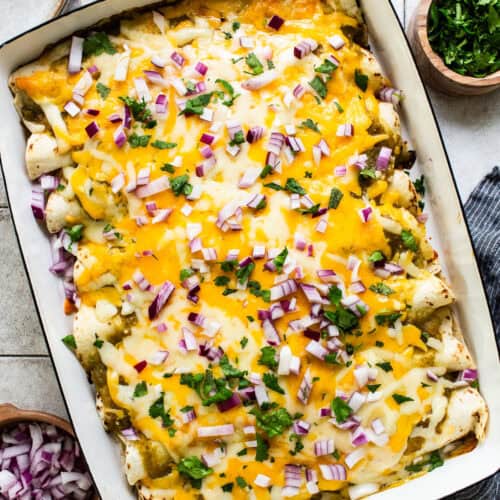 Spinach enchiladas in a baking dish topped with melted cheese, salsa verde, onions, cilantro, and sour cream or Mexican crema.