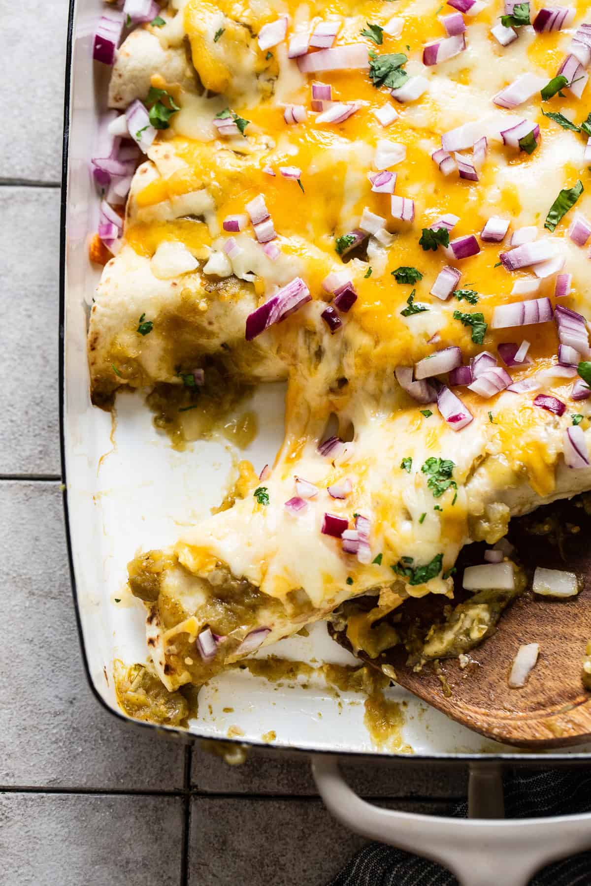 A wooden spatula grabbing a spinach enchilada from a baking dish to serve it.