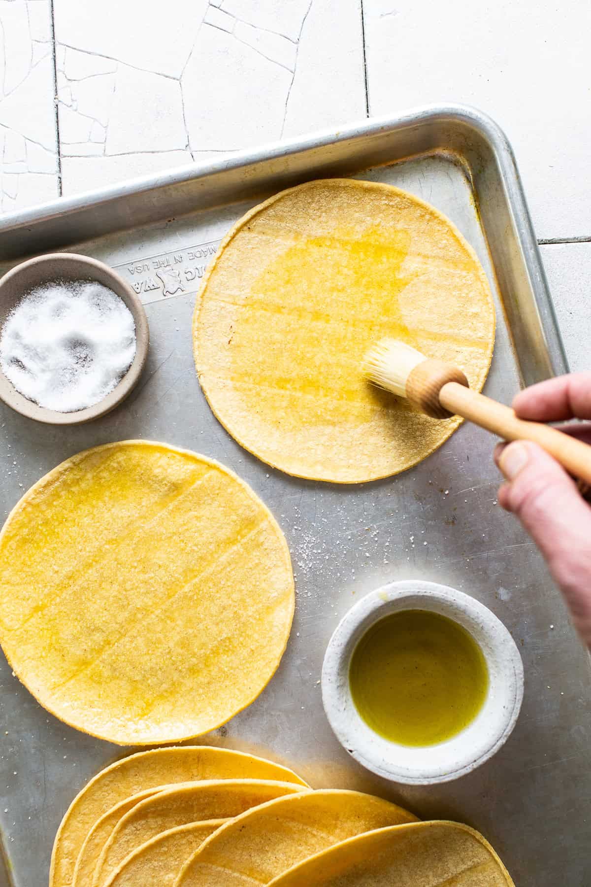 Brushing oil on both sides of a corn tortilla on top of a baking sheet.