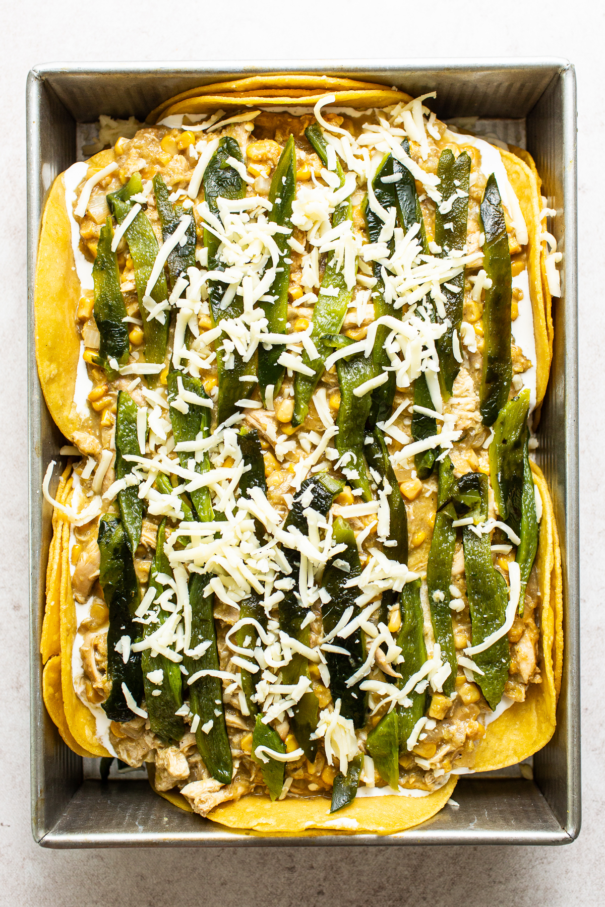 A layer of corn tortillas in a baking sheet topped with Mexican crema, chicken and corn mixture, roasted poblanos, and cheese.