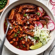 A large bowl of pozole rojo served with limes, oregano, radish slices, cilantro, and cabbage.