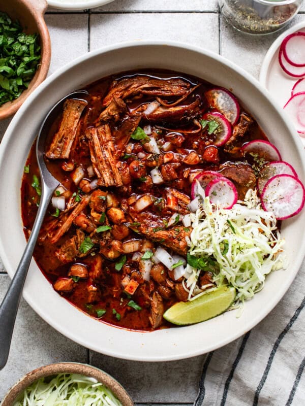 A large bowl of pozole rojo served with limes, oregano, radish slices, cilantro, and cabbage.