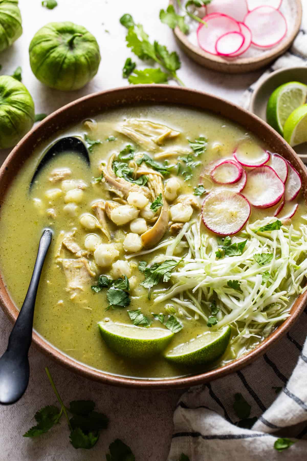 A warm bowl of homemade green chicken pozole verde topped with thinly sliced radishes, chopped cilantro, lime wedges, and shredded cabbage.