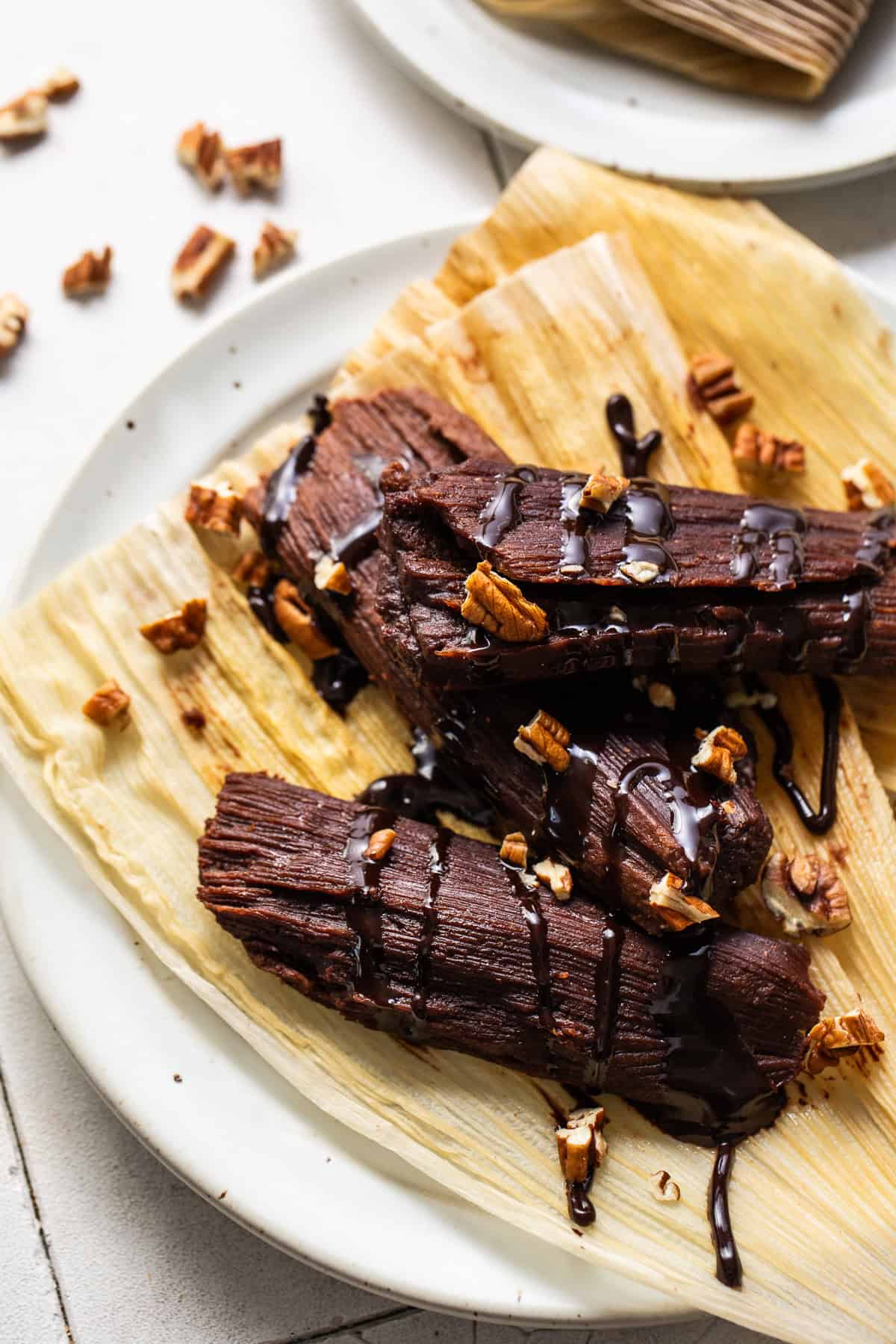 Three chocolate tamales on a plate drizzled with a little chocolate sauce and garnished with some chopped pecans.