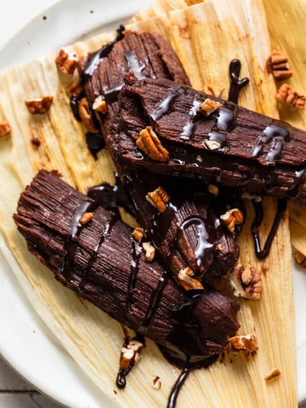 Chocolate tamales drizzled with a chocolate sauce and chopped pecans on a corn husk.