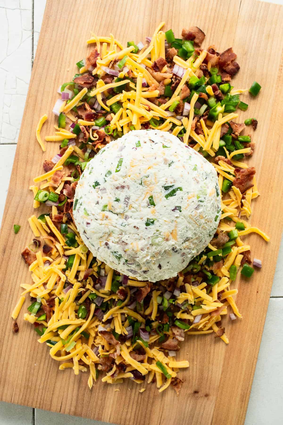 A formed cheese ball placed on top of a bed of shredded cheese, diced bacon, jalapeños, red onion and green onion before being rolled and served.