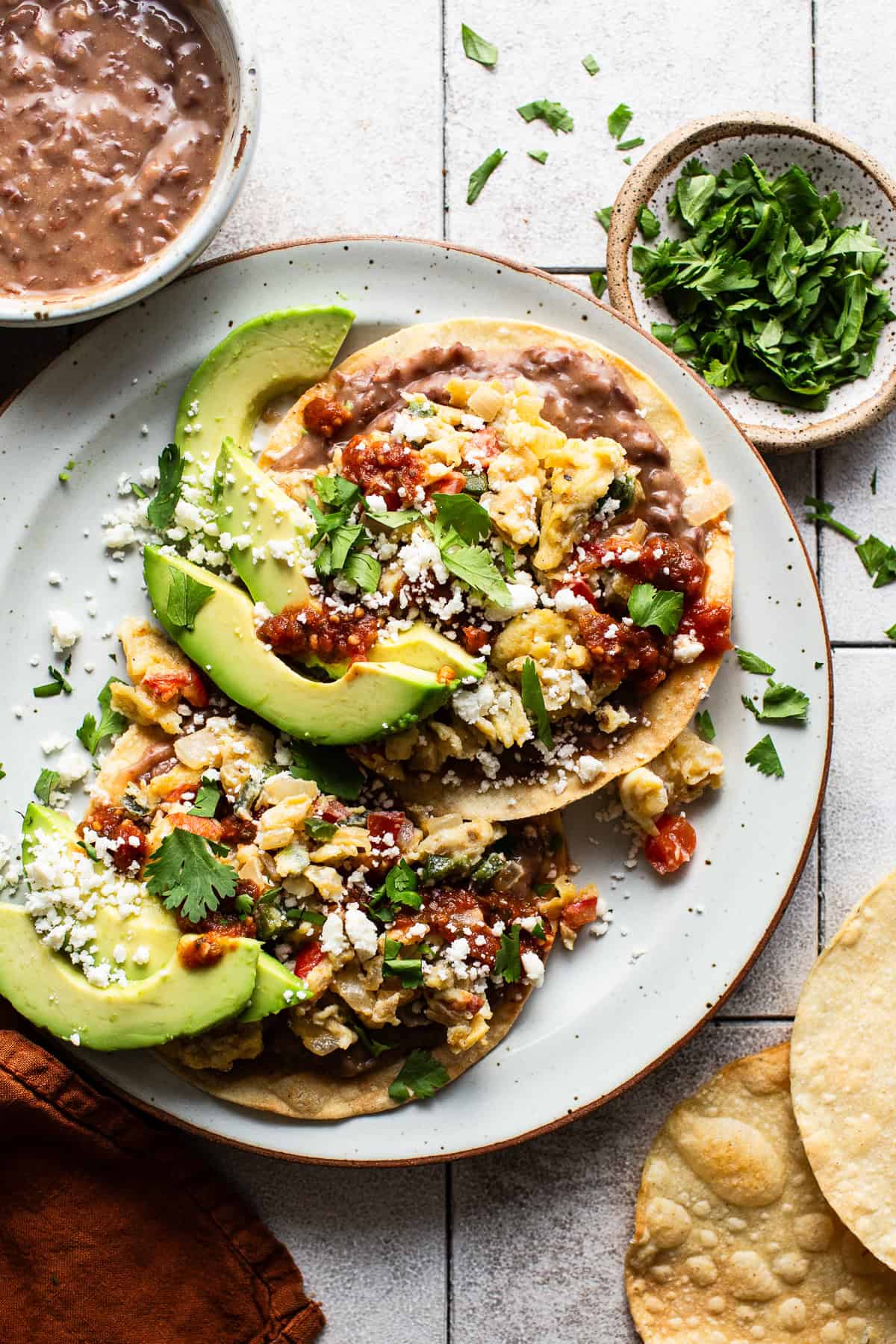 A breakfast tostada assembled on a plate with beans, scrambled eggs, salsa, avocado, cotija cheese, and cilantro on top.