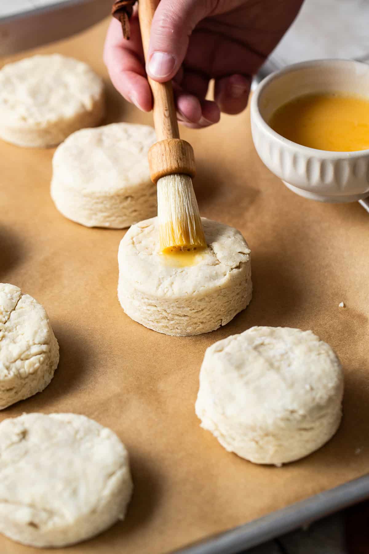 Masa harina biscuits on a sheet pan lined with parchment paper being brushed on top with an egg wash before being baked.