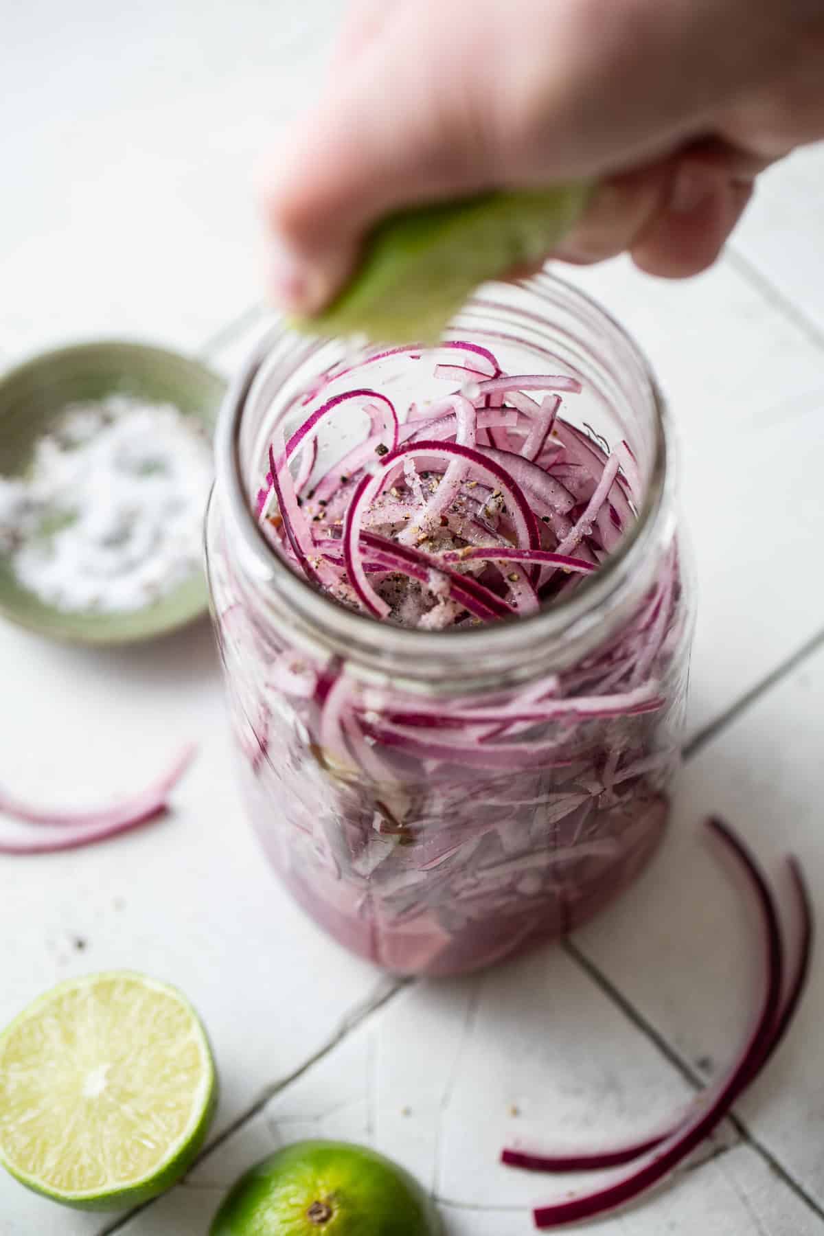 Fresh lime juice being squeezed into a jar of thinly sliced red onions seasoned with kosher salt and black pepper.