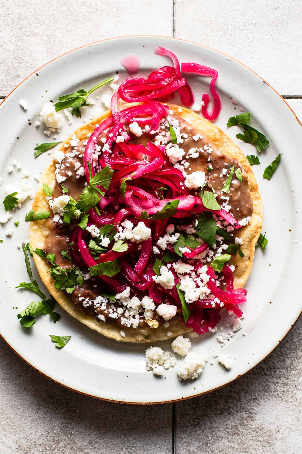 A tostada topped with bright mexican pickled onions, cotija cheese, cilantro, and beans.